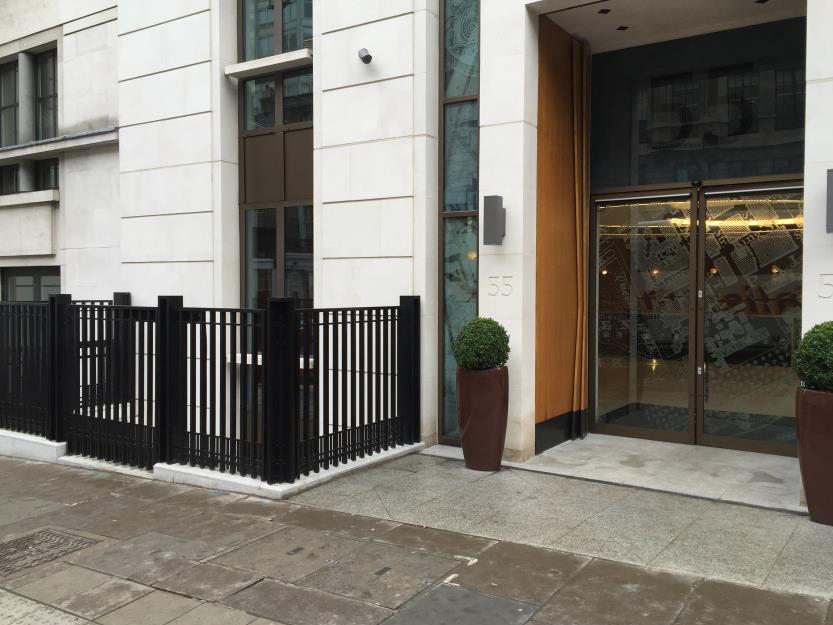 Modern building entrance with a lightwell enclosed by black metal railings and decorative planters on either side of the glass doors.