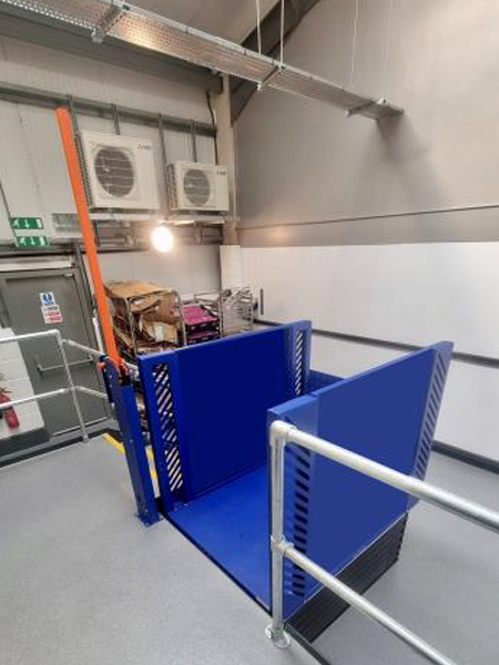 Blue industrial goods lift installed in a modern facility with safety railings and visible air conditioning units in the background.