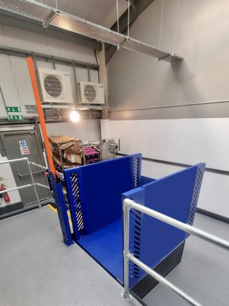Blue industrial goods lift installed in a modern facility with safety railings and visible air conditioning units in the background.