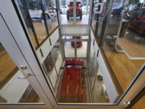 Red car being lowered in a glass-enclosed goods lift within a modern showroom setting.