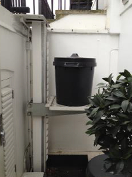Compact lightwell lift holding a black bin, installed in a narrow outdoor space with white walls.